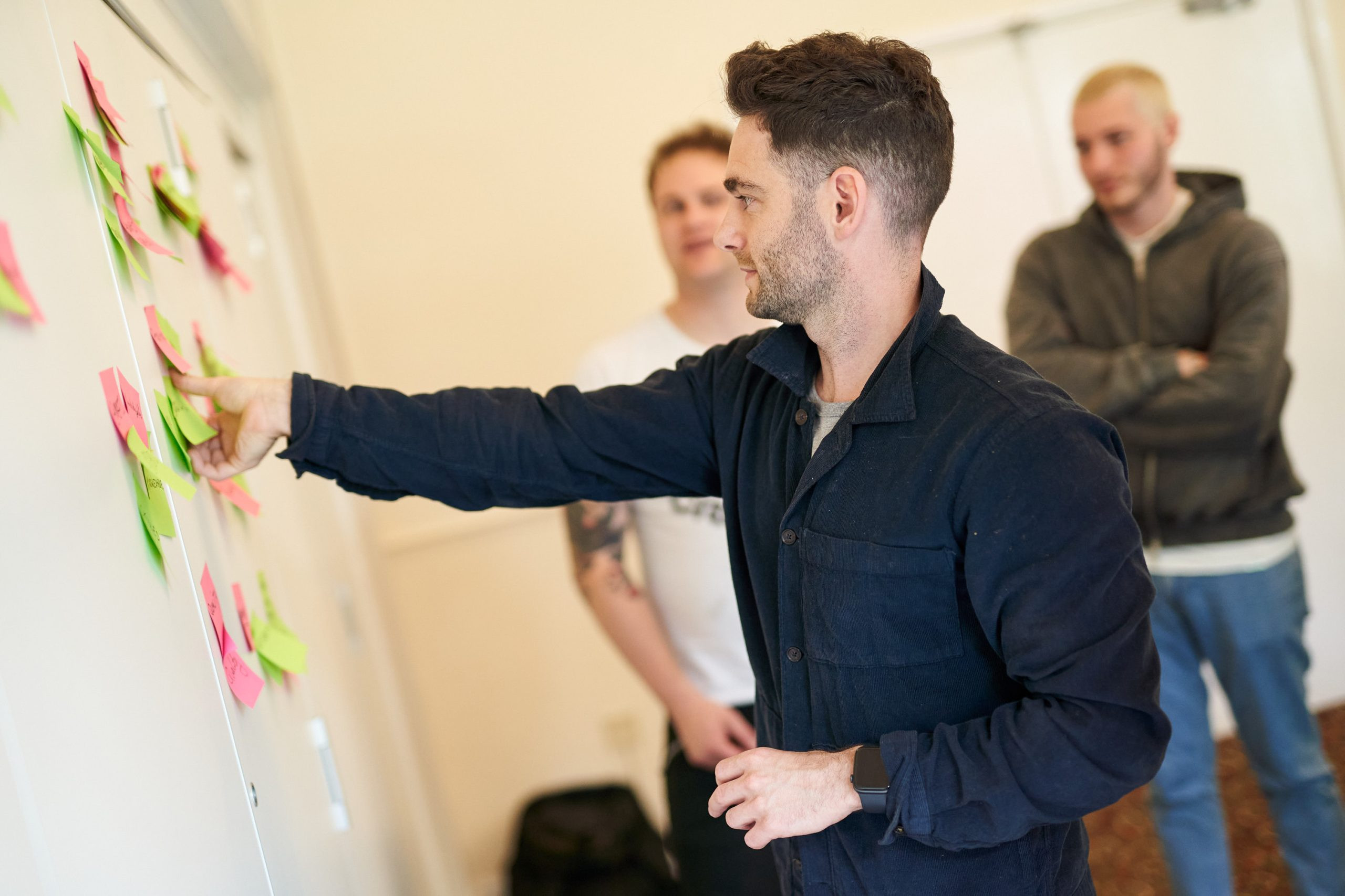 Three men standing in front of giant whiteboard covered in post it notes. One man sticks post it to wall.